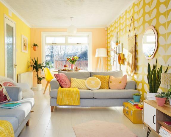 Giocoso soggiorno giallo e grigio con pittura murale gialla, carta da parati a motivi retrò e divani grigio chiaro in stile con cuscini assortiti.