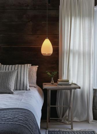 Dormitor in stil rustic cu perete din lemn, perdea de in, lumina suspendata deasupra noptierei, lenjerie de pat texturata, covor