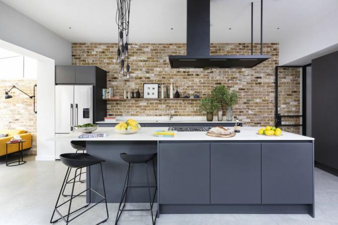zwarte keukenunits met witte werkbladen, grijze tegelvloer, moderne afzuigkap, bakstenen muur, hanglampen, barkrukken, kookeiland met kookplaat en eetbar