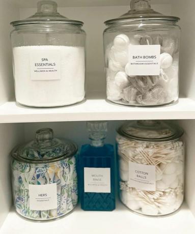 Un armario de baño con frascos de vidrio llenos de elementos esenciales para el baño, como sales de Epsom, bolas y bastoncillos de algodón, tampones, etc.