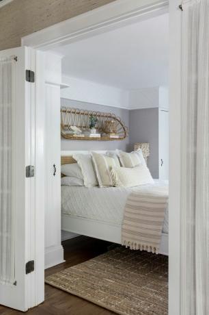 pilkas ir baltas miegamasis su rotango lentyna virš lovos, neutralios tekstūros patalynė, medinės grindys, kilimėlis