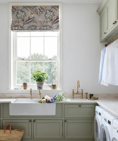 Gedämpfte salbeigrüne Schränke im Waschraum mit Doppelwaschbecken, Messinghähnen, weißen Wänden und geblümter Fensterjalousie