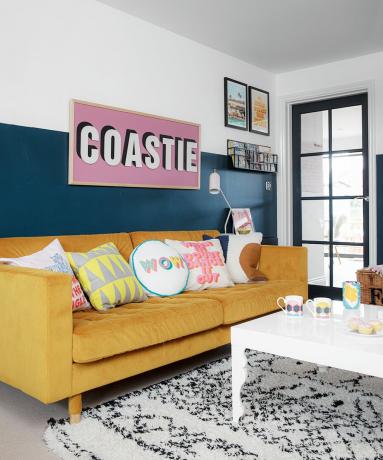 Gul sofa med diverse, lekne puter, tofarget malt vegg, taktilmønstret monoteppe og stort skrivefeilskilt på veggen.