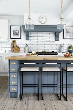Cozinha com unidades brancas estilo Shaker, ilha azul, bancos altos de metal preto e luminárias pendentes de vidro
