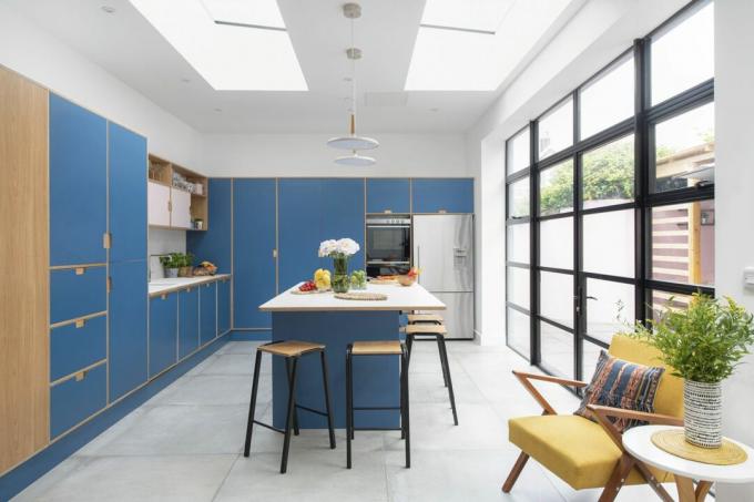 Plano general de la cocina con baldosas grises de gran formato, unidades de madera contrachapada de fórmica azul y rosa, encimera blanca y puertas al jardín estilo Crittall