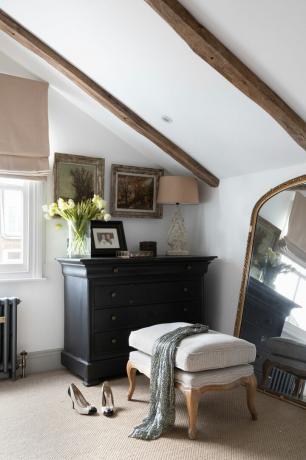 ピッパジョーンズの家：ダークブルーブラックの塗装ドレッサー、ベージュのオットマン、大きな金枠の鏡、ベージュのカーペット、木製の梁のある白い傾斜天井のある予備の寝室の隅