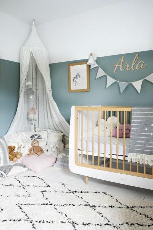 Chambre de bébé avec murs peints en bleu, tapis monochrome, lit bébé blanc et chêne et baldaquin blanc