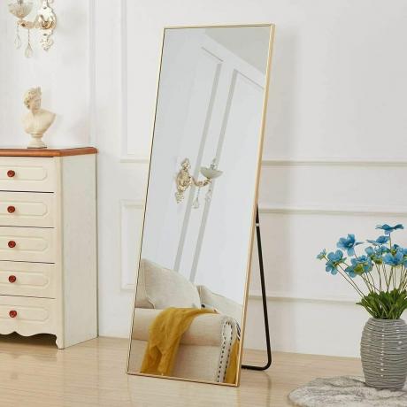 Arany teljes hosszúságú álló tükör egy szobában mellszoborral, kanapéval és virágokkal