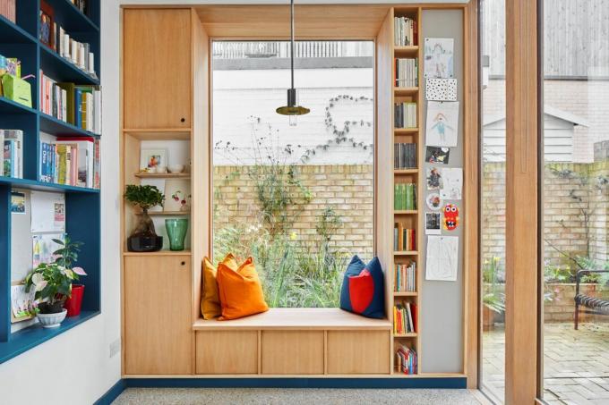 Asiento de ventana de madera incorporado con cojines, rodeado de estanterías para libros de cocina
