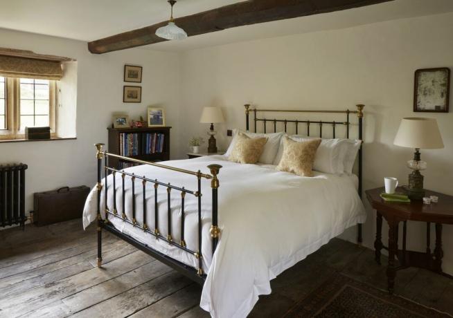 Cama de hierro con ropa de cama blanca y almohadas doradas en el dormitorio con paredes de color crema y suelos de madera.