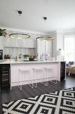 Cozinha com móveis de parede com efeito de concreto, ilha rosa claro, banquetas altas de metal branco, piso de madeira escura, tapete geométrico monocromático e pendentes circulares marcantes