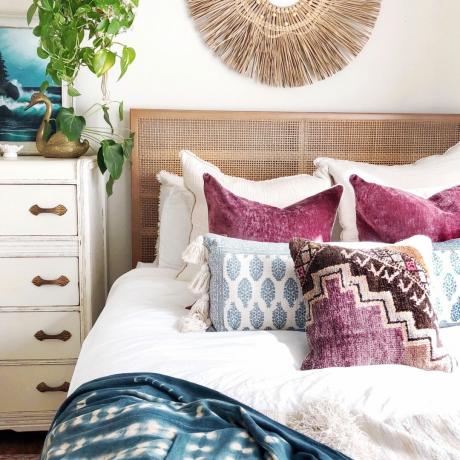 Farverig boho-inspireret seng med sengegavl i sukkerrør, vægstykke med naturlig tekstur, efterstillede stueplante og diverse mønstrede og bloktrykte spredepuder i blomme og blå nuancer.