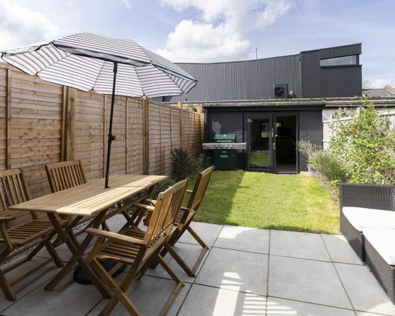 Een externe opname van achtertuin met tuinbijgebouw, houten tuintafel en -stoelen en parasol met gestreept decor