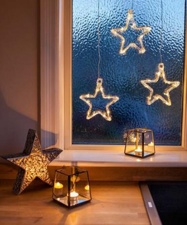 En trio batteridrevne stjerneformede julelys på vinduet med to sorte innrammede lykter med telys og metallisk stjernedekor