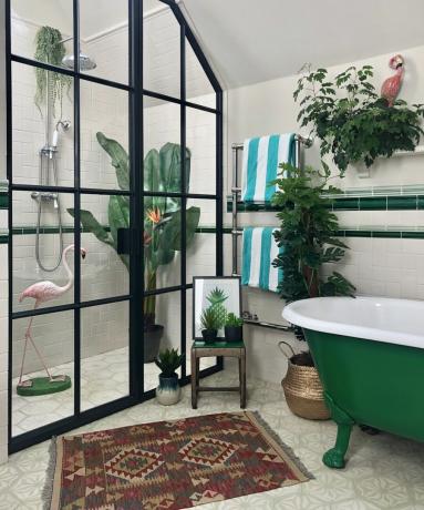 Kylpyhuone, jossa on crittal -tyylinen musta kehystetty suihku, tummanvihreä vapaasti seisova kylpyamme, atsteekkimatto ja kasveja