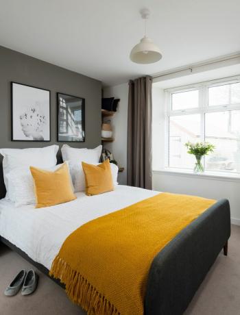 Главная спальня в серой цветовой гамме с двуспальной кроватью, горчично-желтым покрывалом и стеллажом в углу комнаты.