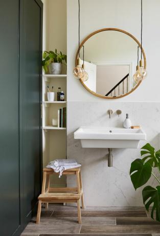 Riippuvalaisimet: Broch. Kylpyhuone, jossa on marmorivaikutteiset laatat seinän puolivälissä, pyöreä peili, joka on kehystetty molemmin puolin paljailla polttimoilla ja riippuvalaisimet