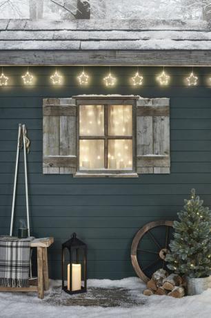 क्रिसमस खिड़की प्रदर्शित करता है: पर्दे की रोशनी, मोमबत्तियों और उत्सव रोशनी के साथ उत्सव हरा शीतकालीन केबिन