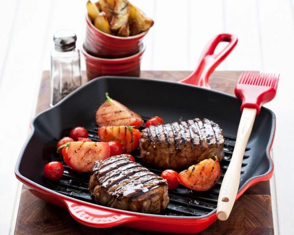 Una plancha roja Le Creuset con bistec y tomates asados, con cepillo de silicona y cazuelas con gajos de patata
