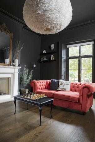 Musta olohuone puulattialla, punainen Chesterfield sohva, musta sohvapöytä shakkilaudalla ja ylisuuri höyhenkattovalo
