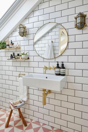 Ubin metro putih di kamar mandi dengan ubin lantai merah muda dan putih, sconce dinding industri, dan faucet/pipa emas