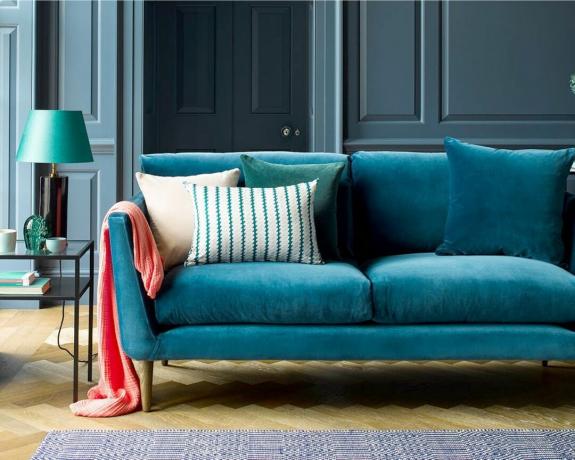 Un sofá de terciopelo azul turquesa con patas de madera en una sala de estar con paredes con paneles pintados de azul
