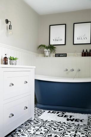ห้องน้ำสีครีมที่มีผนังกรุ ปูพื้นด้วยกระเบื้องลวดลายขาวดำ อ่างอาบน้ำม้วนด้านบนสีน้ำเงิน และโต๊ะเครื่องแป้งสีเทาสไตล์เชคเกอร์