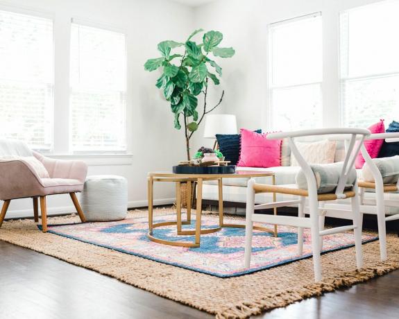 Jasny i przewiewny salon boho z warstwowymi dywanikami, dużą rośliną w rogu, zakrzywionym stolikiem kawowym i różowymi popowymi poduszkami na sofie.