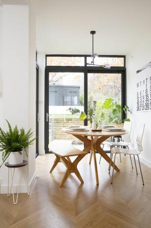 Área de comedor de una cocina de planta abierta en la casa de María con mesa y banco de madera, sillas blancas y puertas corredizas con marco negro