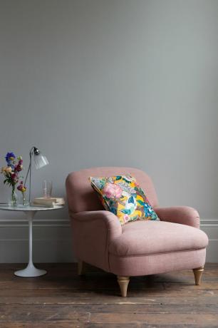 różowe krzesło w pokoju z drewnianą podłogą, szarymi ścianami i bocznym stolikiem ze świecącymi, książkami i lampą