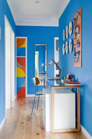 Modře vymalovaná chodba se psacím stolem a židlí, deskami na zdi a na konci dveřmi skříně pomalovanými zakřivenými geometrickými vzory v červené, oranžové, žluté a modré barvě