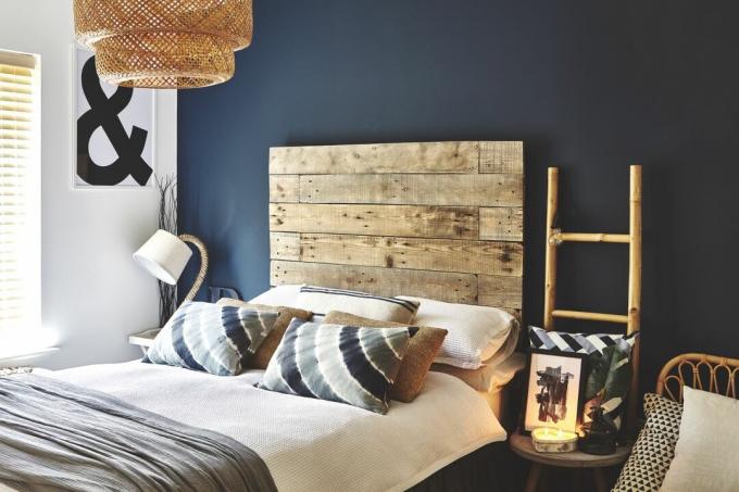 Ersatz-Schlafzimmer mit übergroßem Holzpaletten-Kopfteil gegen eine dunkelblaue Wand, Leiterregale und Rattan-Pendelleuchte