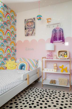 Chambre d'enfant avec papier peint arc-en-ciel sur un mur, effet de peinture festonnée rose sur l'autre. Lit blanc avec coussins arc-en-ciel et soleil, tapis à pois noir et blanc, étagères ouvertes Scandi et imprimé astronaute sur le lit
