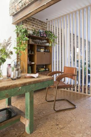 Essbereich mit Korkboden, freiliegender Holzwand, Raumteiler aus Lattenrost, der zum Wohnzimmer führt, und altem Holztisch mit grün lackierten Beinen