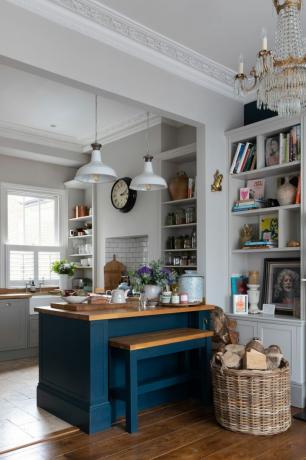 Rumah Pippa Jones: bidikan ke arah dapur dengan semenanjung dicat biru dan bangku built-in, keranjang anyaman dengan kayu gelondongan, dan lampu gantung putih di atas kepala