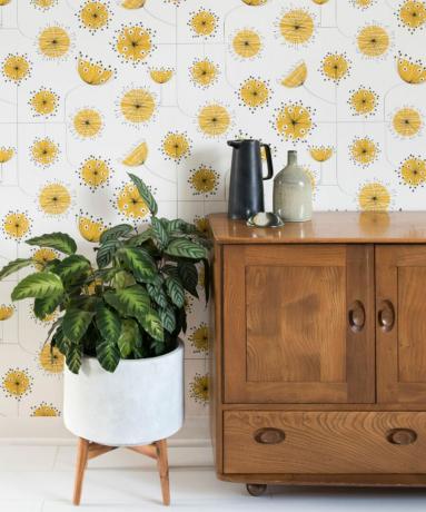 Leuk retro bloemenbehang in wit en mosterdkleur, achter een potplant op pootjes en een houten dressoir.