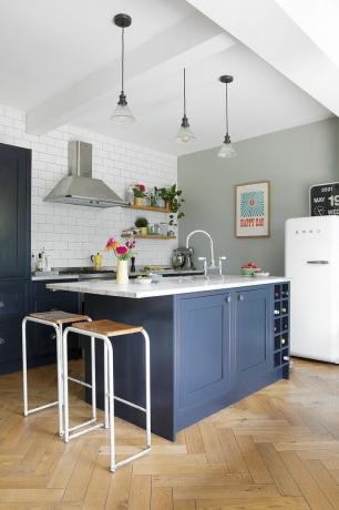 Kökets hörn med blå Shaker-stil, barstolar i trä och vit metall, fiskbensgolv, glashängen ovanför och ljusgrön bakvägg