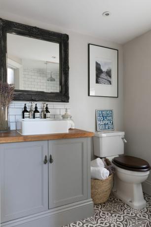 Casa de Pippa Jones: banheiro com pia cinza, espelho de bronze, piso com padrão monocromático e impressão " Nunca é tarde para viver felizes para sempre"
