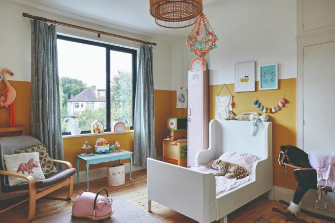 Lea-Wilson hus: børneværelse med gule og hvide farveblokvægge, hvid seng med høje sider, grå lænestol, blåt skrivebord og loftlampe i rotting