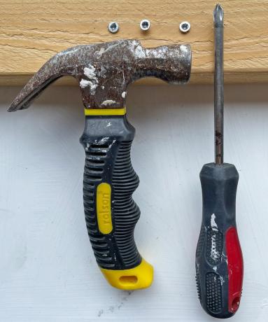 Beschädigte Schrauben in einem Stück Holz, daneben ein Schraubenzieher und ein Hammer