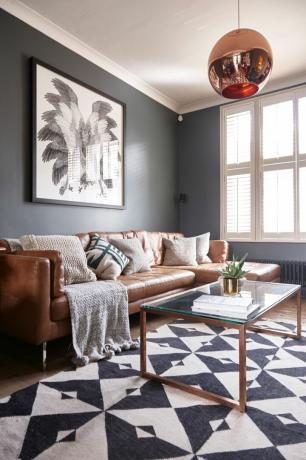 Jasono Traveso namas: svetainė su tamsiai pilkomis sienomis, įdegusios odos sofa, minimalistinis kavos staliukas su stikliniu viršumi, varinė pakabinama šviesa ir nespalvoto rašto kilimas virš medinių grindų