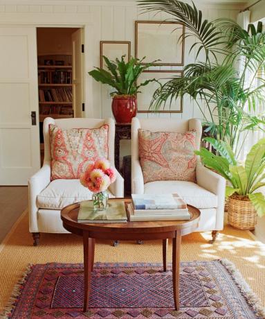 Dvě bílá čalouněná křesla s korály, vzorované zadní polštáře ve velkém obývacím pokoji s kulatým dřevěným konferenčním stolkem, vzorovaným kobercem a spoustou pokojových rostlin