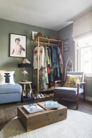 Kotitoimiston alue, jossa avoin vaatekaappi täynnä vaatteita ja kenkiä, oleskelutila sohvalla ja nojatuoli, sohvapöytä, jonka päällä on muotilehtiä ja kehystetty valokuva oliivinvihreää vasten seinään