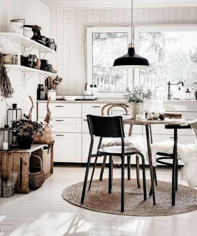 Nápad na stolovanie pre malé kuchyne s bielym obložením stien, drevenou podlahou a bielymi policami s malým okrúhlym stolíkom, nízkym čiernym závesným svetlom a jutovým kobercom