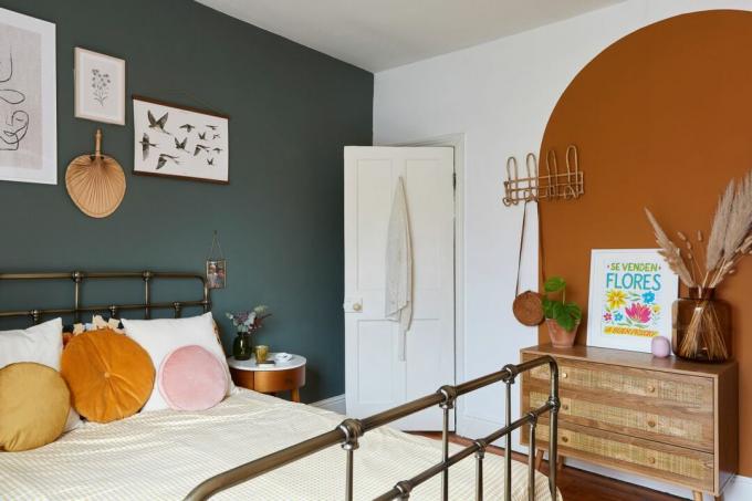 ห้องนอนใหญ่ที่มีผนังสีเข้มด้านหนึ่งและซุ้มประตูสีส้มพร้อมเฟอร์นิเจอร์หวายและเตียงโลหะสีดำ