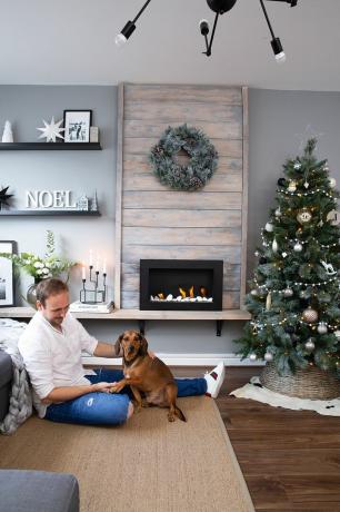 JT, ahşap panelli şöminesi ve beyaz ve gri süslemeli Noel ağacı olan gri oturma odasında köpek Rocco ile oynuyor.