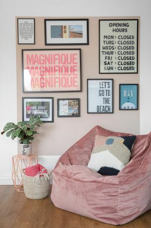 ركن من غرفة ذات مخطط مفتوح مع كرسي من القماش المخملي الوردي ، مربع مطلي باللون الوردي على الحائط مليء بالمطبوعات لإنشاء جدار معرض