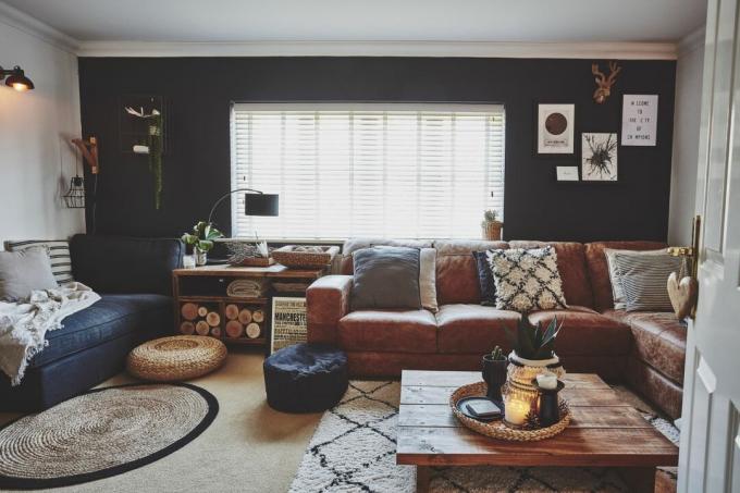 Stue med en mørk svart/blå vegg, andre hvite vegger, L-skinnsofa i brun skinn, sofa i svart stoff, tepper, rottingpuffer og salongbord laget av palettplater i tre