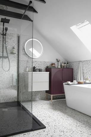 Kamar mandi dengan dinding teraso dan ubin lantai, bilik pancuran berbingkai hitam, bak mandi putih, meja rias gantung abu-abu, cermin bundar, dan kabinet ungu