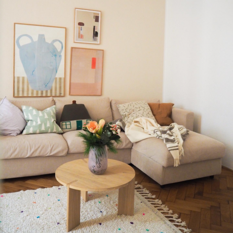 Glad, lekfull loungevrå med abstrakt, pastellfärgad väggkonst, schäslong med diverse, mönstrade strökuddar och litet runt soffbord i trä med vas med färska blommor.
